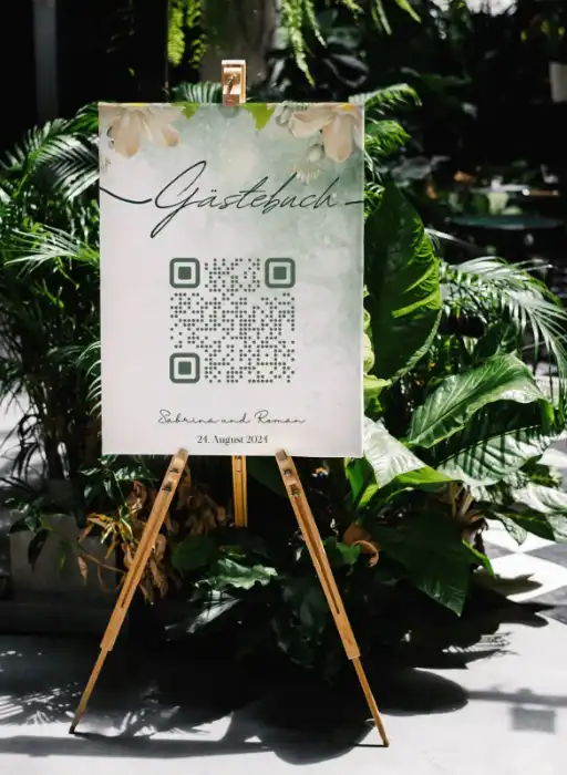 Wedding guest book sign with QR code to collect wedding photos, adorned with plants, labeled Gästebuch for guests.