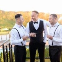 Front view of smiling groom wearing in black suit and bowtie standing with groomsmen on balcony, with view on park, holding glasses with alcohol and celebrating wedding day